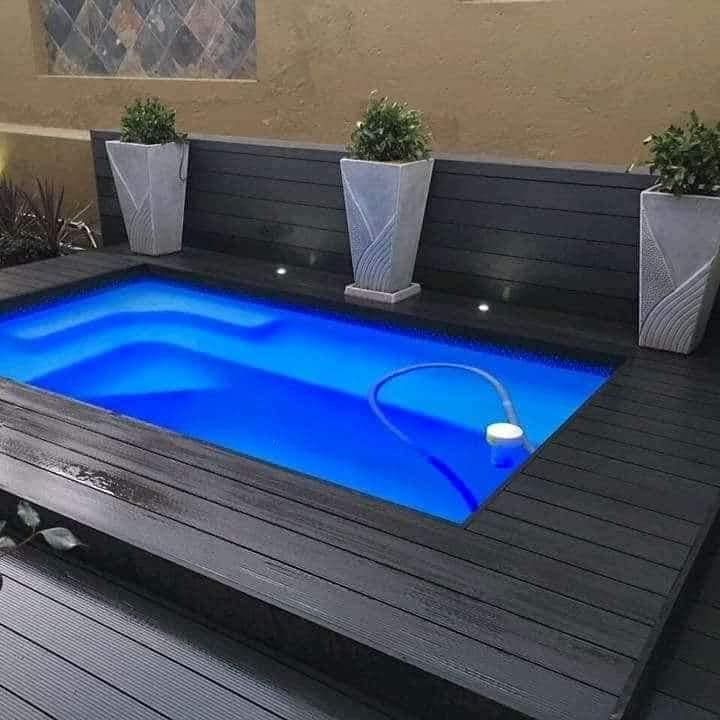 Small rectangular pool with blue water surrounded by a dark gray deck, potted plants, and a dark wall.