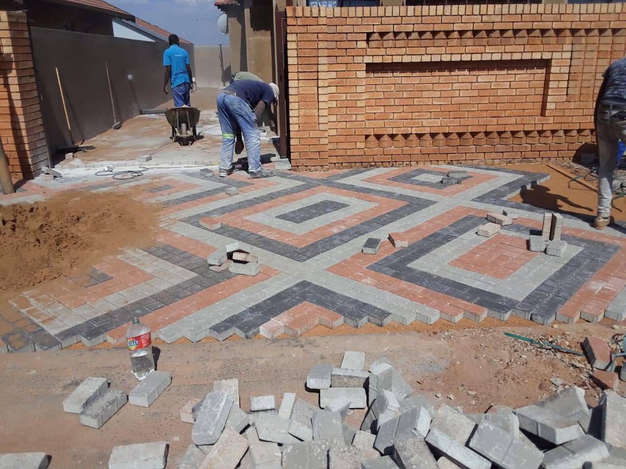 Bricklayers constructing a patterned driveway with gray, orange, and black bricks. Two workers are present.