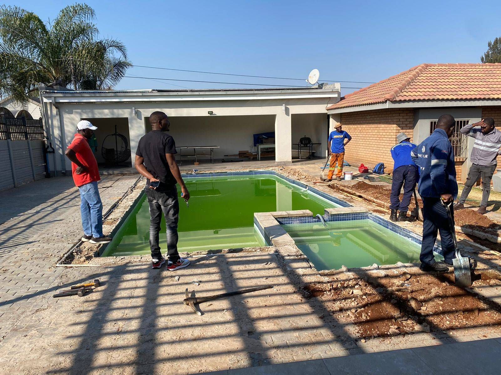 Pool being renovated. Green water in the pool. Workers near the pool. Sunny outdoors.
