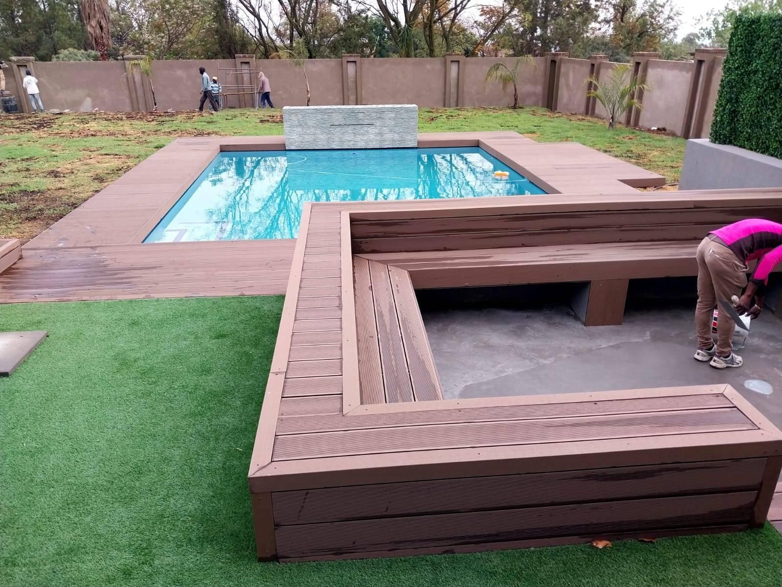 Backyard with pool, brown wooden seating, green lawn, and people working.