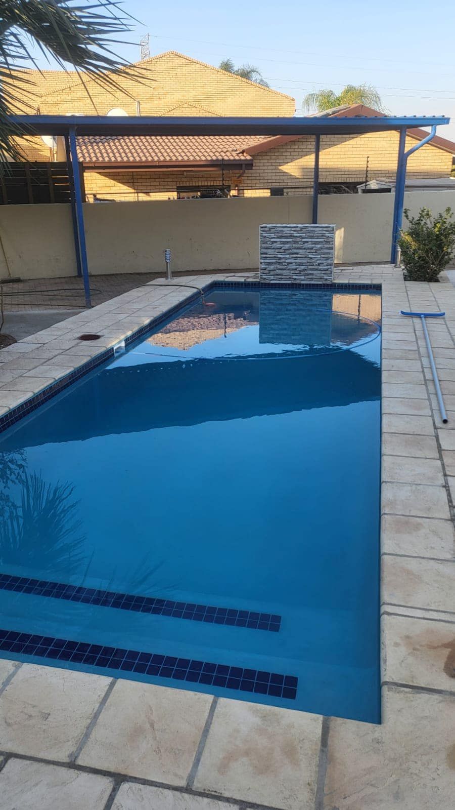 A rectangular blue swimming pool with stone decking, a blue pergola, and a decorative stone feature.