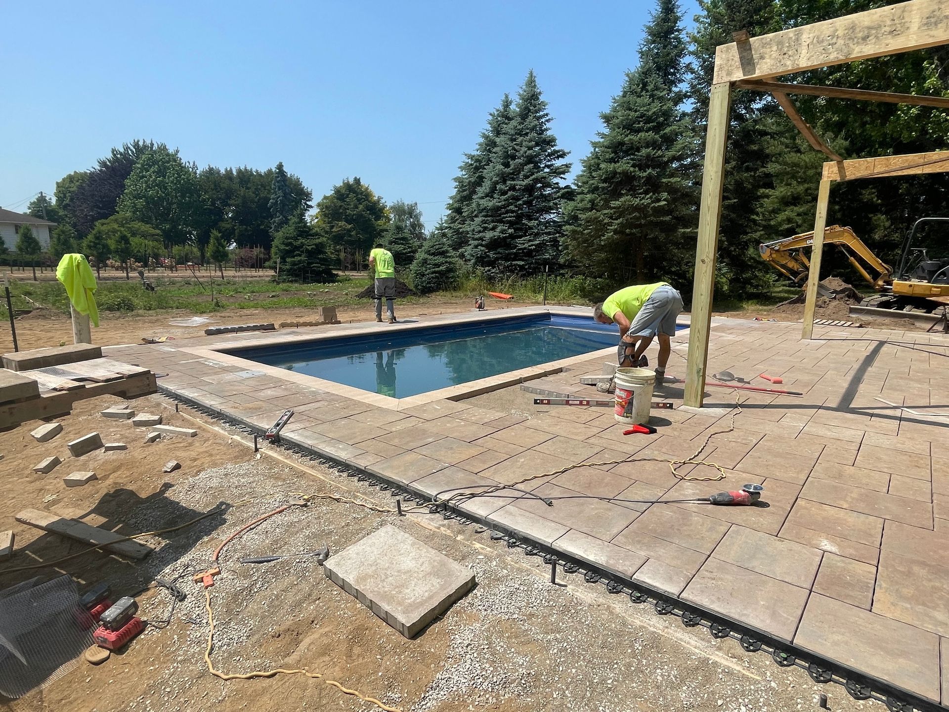 Workers installing pavers around a rectangular swimming pool, under construction. Wooden pergola visible.