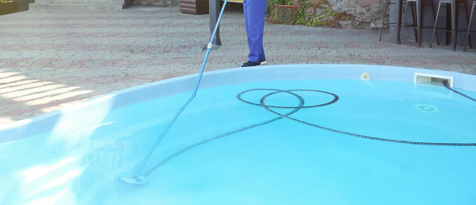 Swimming pool with waterfall feature. Two men near the pool, one standing, one cleaning. Modern buildings in the background.