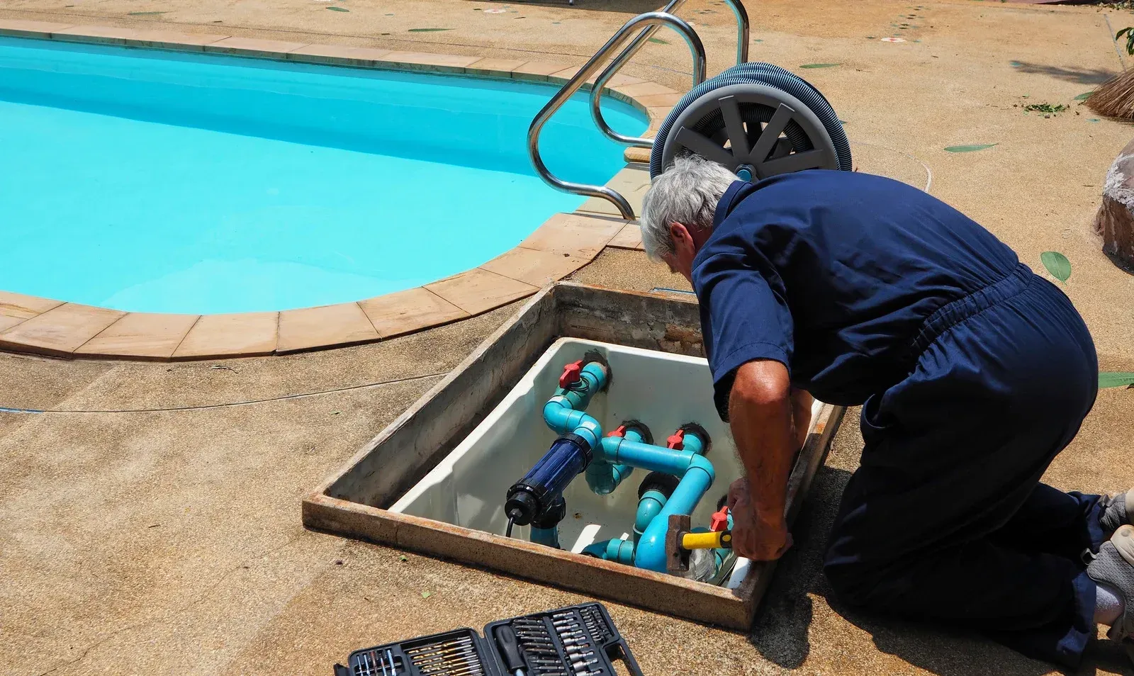 Workers installing patio pavers around a rectangular pool. Sunny day, construction site with trees in background.