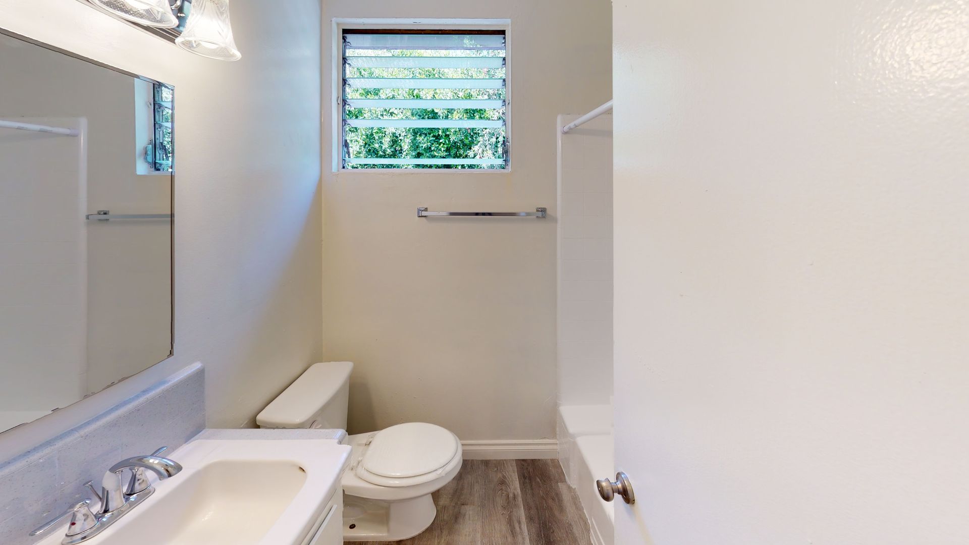 Bathroom with toilet, sink, mirror, and shower, with a window and a towel rack.