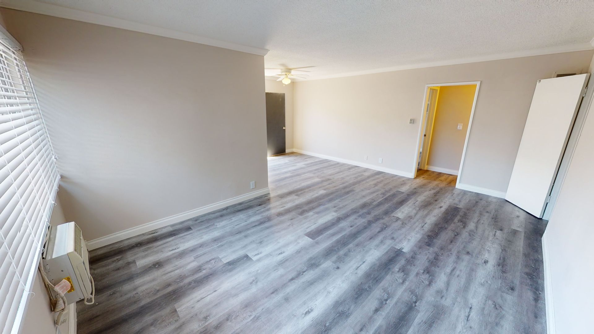 Empty living room with gray wood-look flooring, light walls, and white trim. Window with blinds on the left.