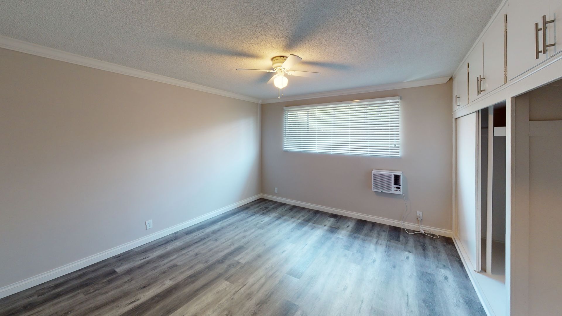 Empty room with gray wood-look floor, window, air conditioner, cabinets, and ceiling fan.
