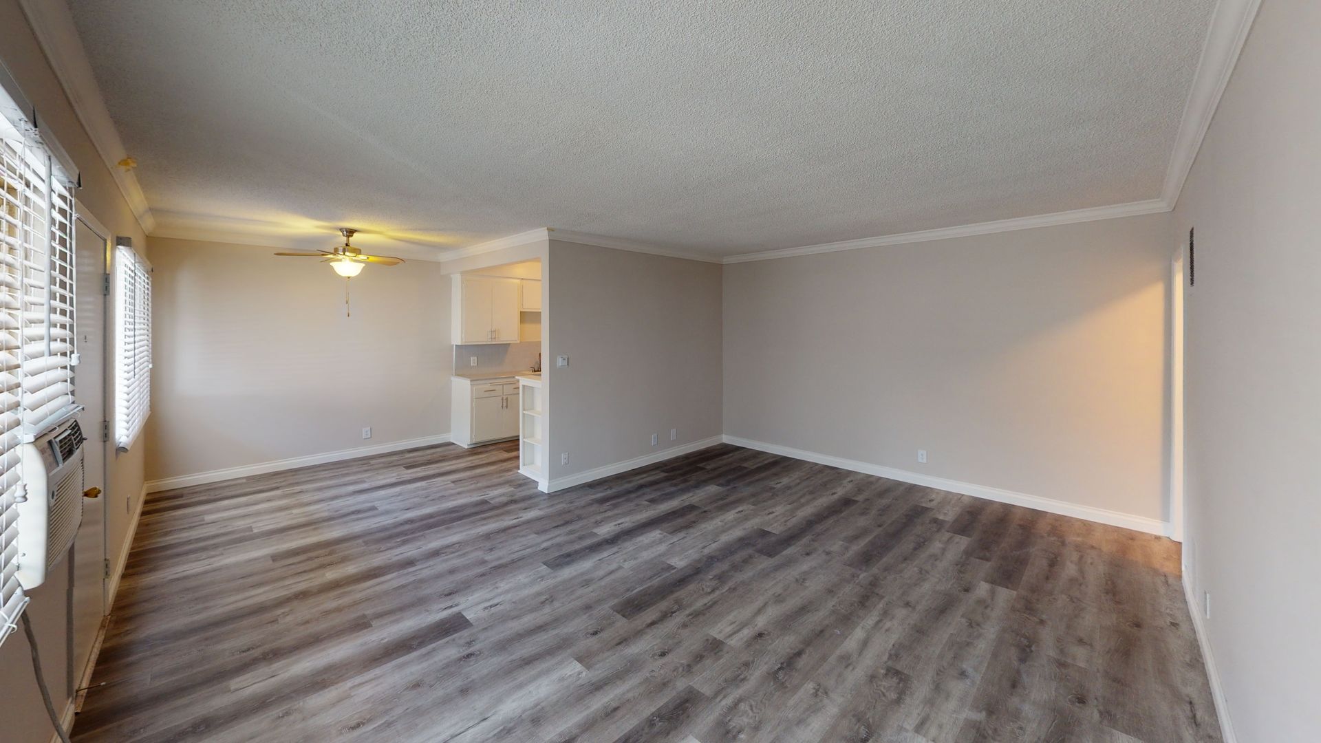 Empty living room with gray wood-look flooring, white walls, and a small kitchen opening.