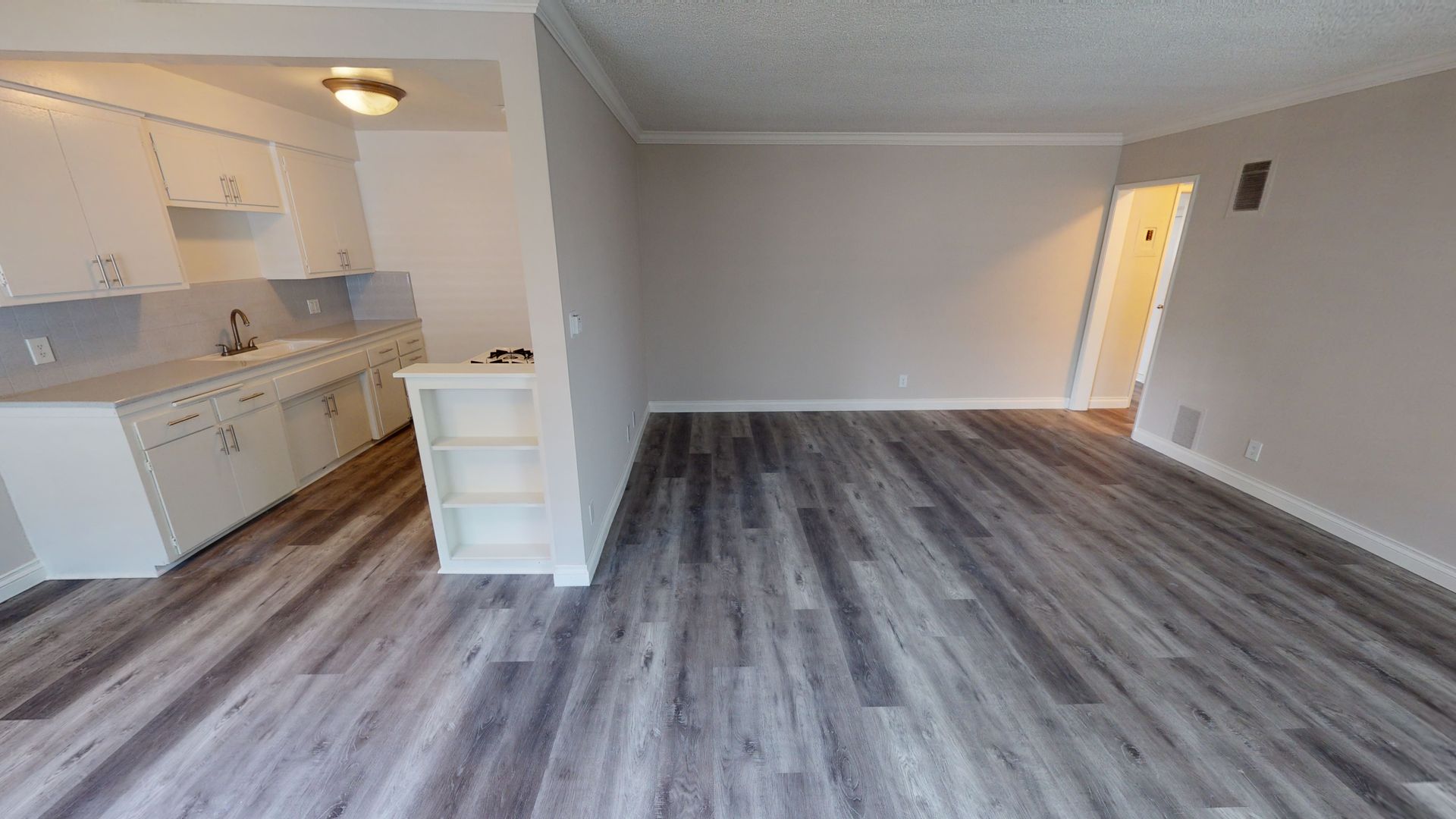 Apartment interior with kitchen and living area, gray wood-look floor, white cabinetry, light gray walls.
