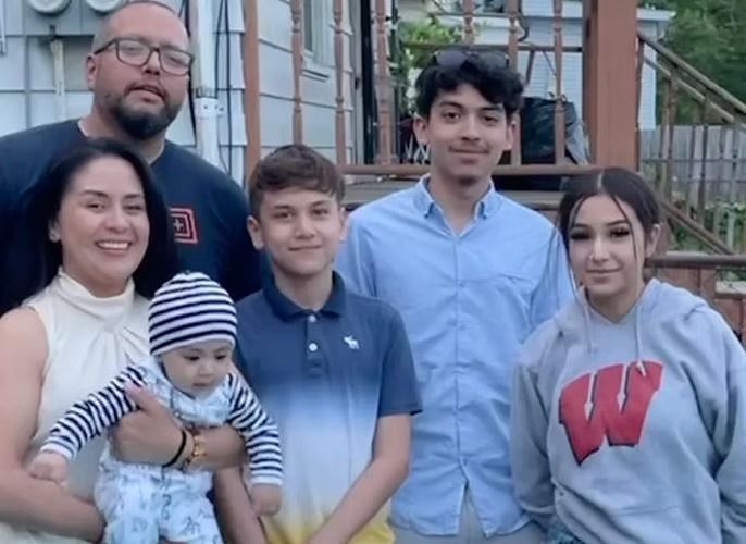 Family portrait: a man with glasses, a woman, and four children posing outside a house. They are smiling.