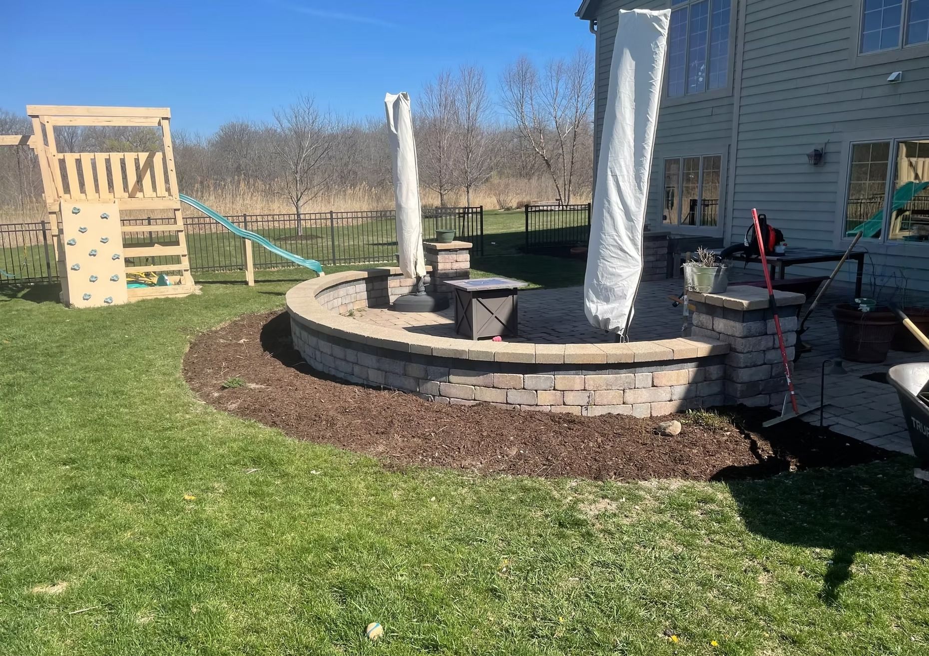 A backyard with a brick fire pit, patio furniture, and a children's playset on a sunny day.  Mulch borders the fire pit.