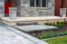 A home's entrance with a stone facade, concrete porch, and steps. A paved walkway leads to the front door with landscaping.