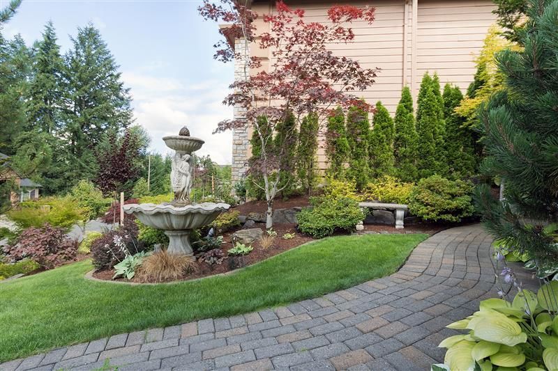 A well-maintained garden featuring a stone fountain, brick pathway, lush greenery, and a bench in front of a house.