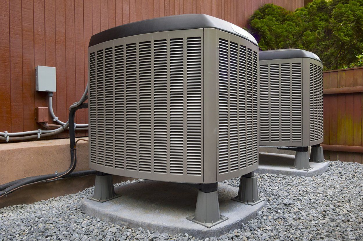 Two outdoor air conditioning units on a gravel base, next to a wooden fence.