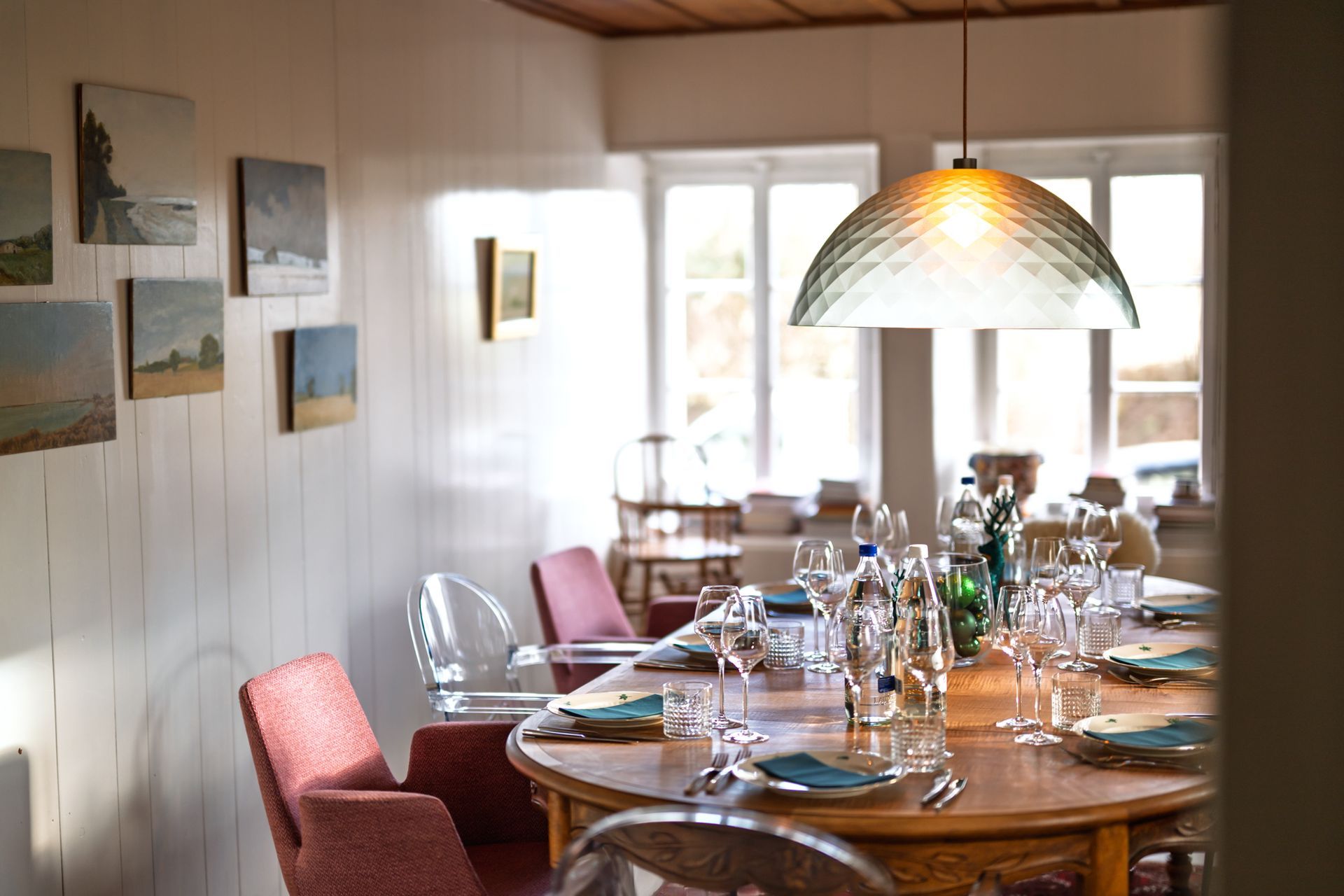 Salle à manger avec table ovale dressée pour le repas, entourée de chaises en velours et transparentes sur un tapis à motifs rouges.