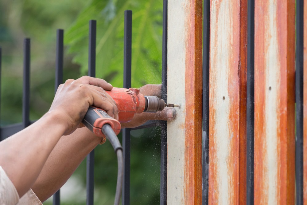 Person's hands using an electric drill to secure wood planks to a fence.