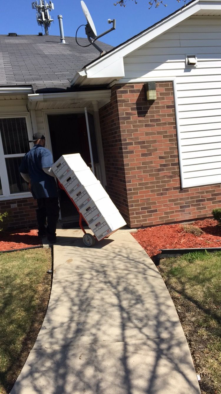 A cardboard box with a tape dispenser and scissors sitting on top, surrounded by other packing boxes on a tiled floor.