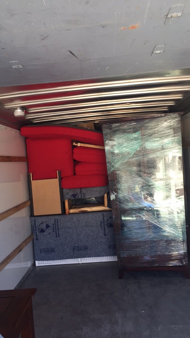 Two workers in blue overalls move a wrapped refrigerator on a dolly and organize boxes in a bright, modern kitchen.