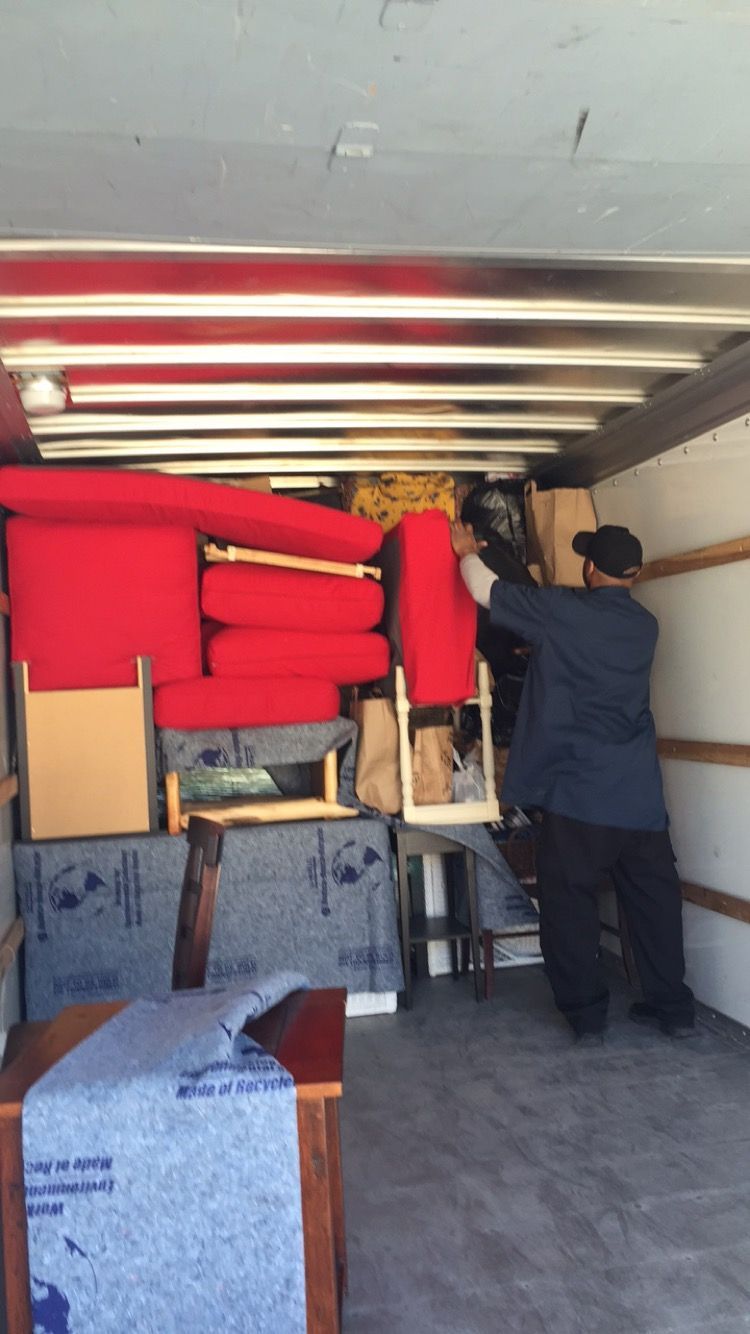 Two workers in blue uniforms unloading cardboard boxes from the back of a white delivery van onto an outdoor sidewalk.
