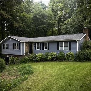 A single-story blue house with black shutters and a brick chimney, surrounded by green lawn and trees.