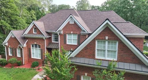 Brick house with brown roof, white trim, and green trees in the background.