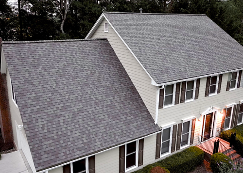 Two-story house with gray shingled roof and light green siding. Dark window shutters, and a welcoming lit entryway.
