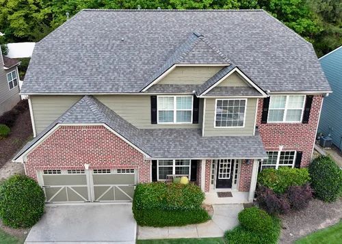 Two-story house with brick and olive green siding, gray roof, and green landscaping. Front door is centered with windows on either side.