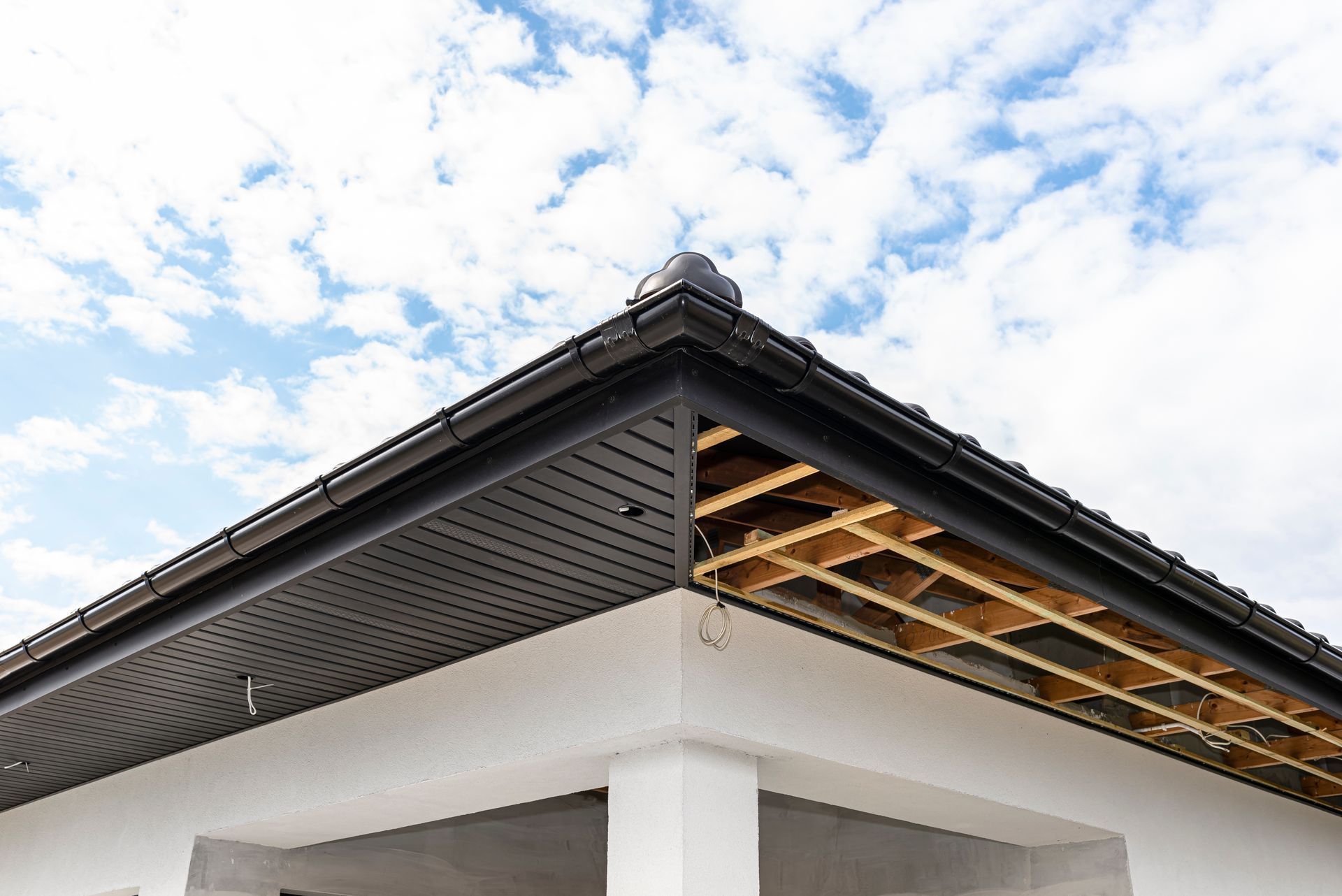 Corner of a white building with a black roof and visible wooden support beams against a cloudy sky.