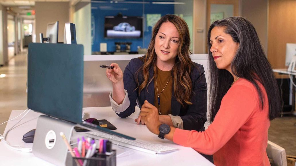 Two women looking at a computer screen, one pointing, discussing work in a modern office.