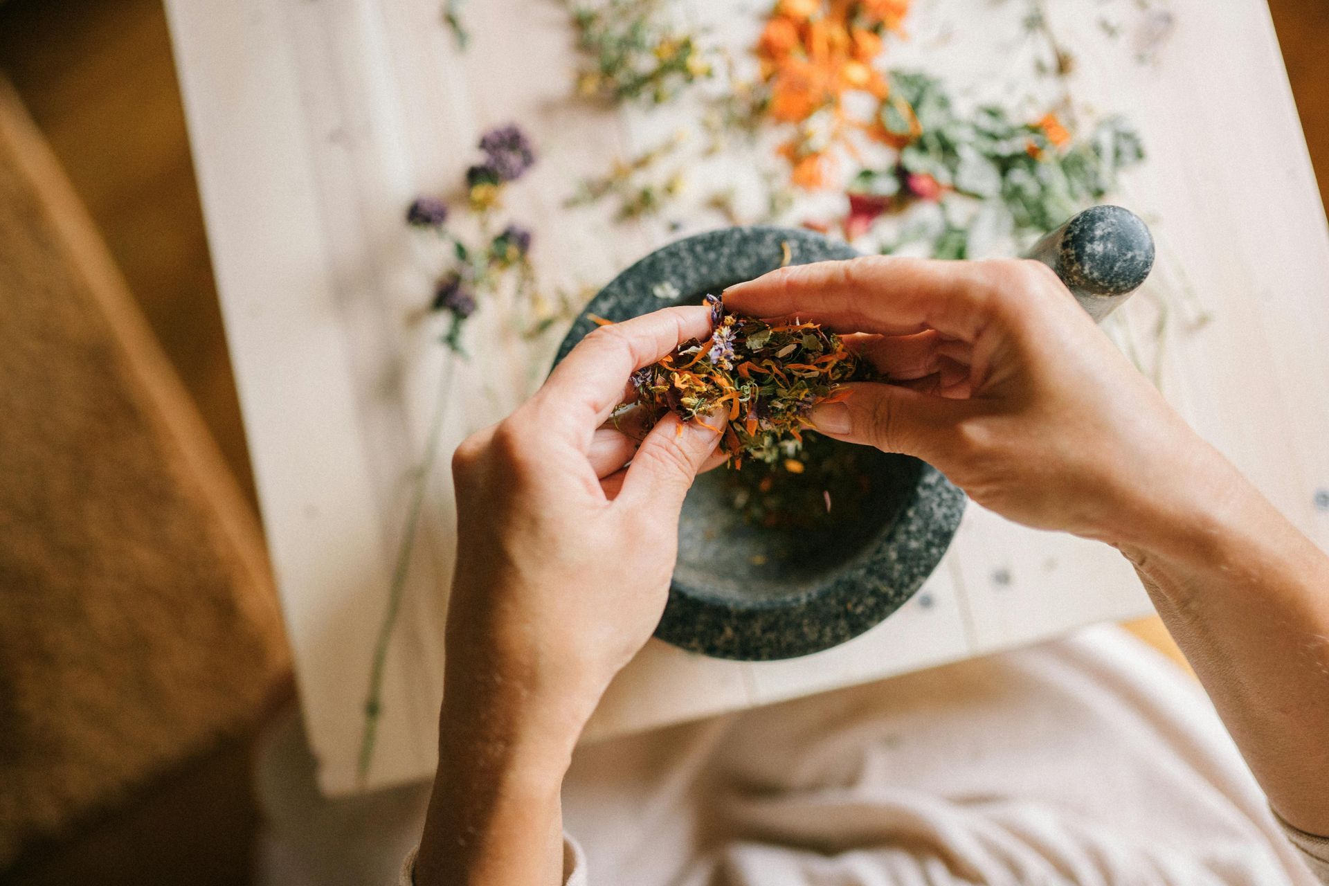 Hands crumbling herbs into a mortar and pestle on a wooden table surrounded by colorful dried flowers.