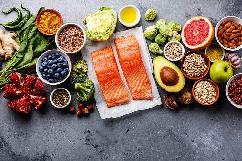 Overhead shot of various healthy foods, including salmon, fruits, vegetables, nuts, and grains, arranged on a gray surface.
