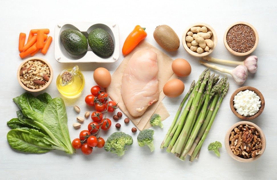 Overhead shot of a white surface with various healthy foods: carrots, avocado, chicken breast, eggs, nuts, vegetables, and fruit.