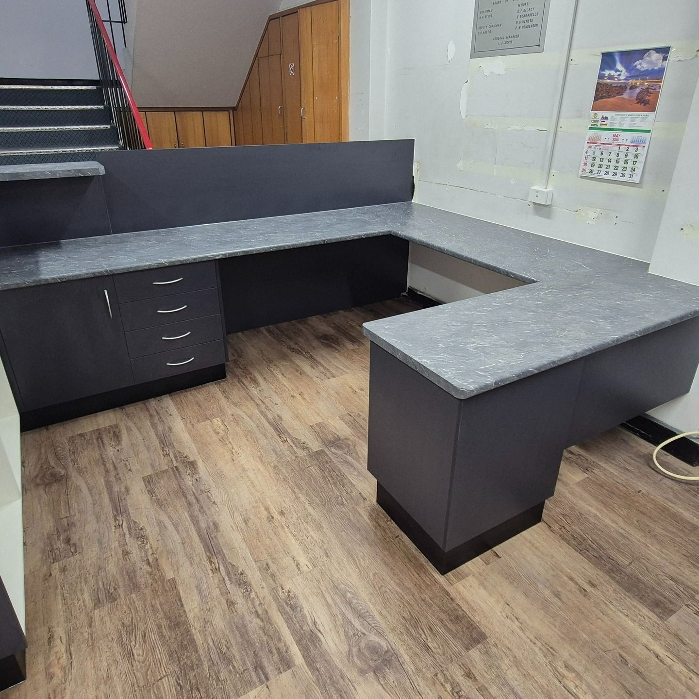 U-shaped Gray Desk With A Cabinet And Drawers On A Wood-look Floor, With Stairs And Cabinets Visible — B & C Cabinets in Mareeba, QLD