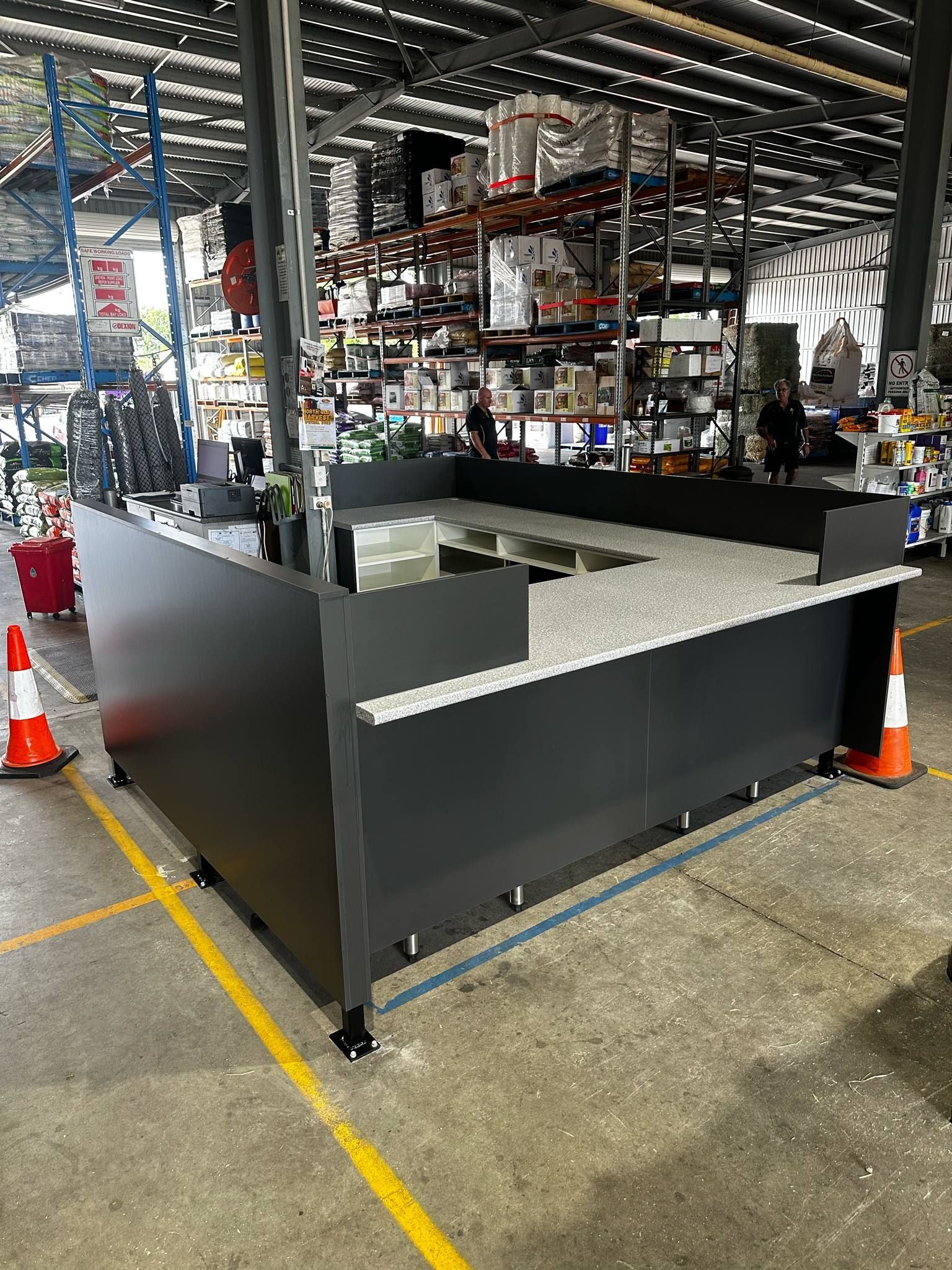 A Dark Gray Retail Counter In A Warehouse Setting, With A Speckled Countertop And Traffic Cones Nearby — B & C Cabinets in Mareeba, QLD