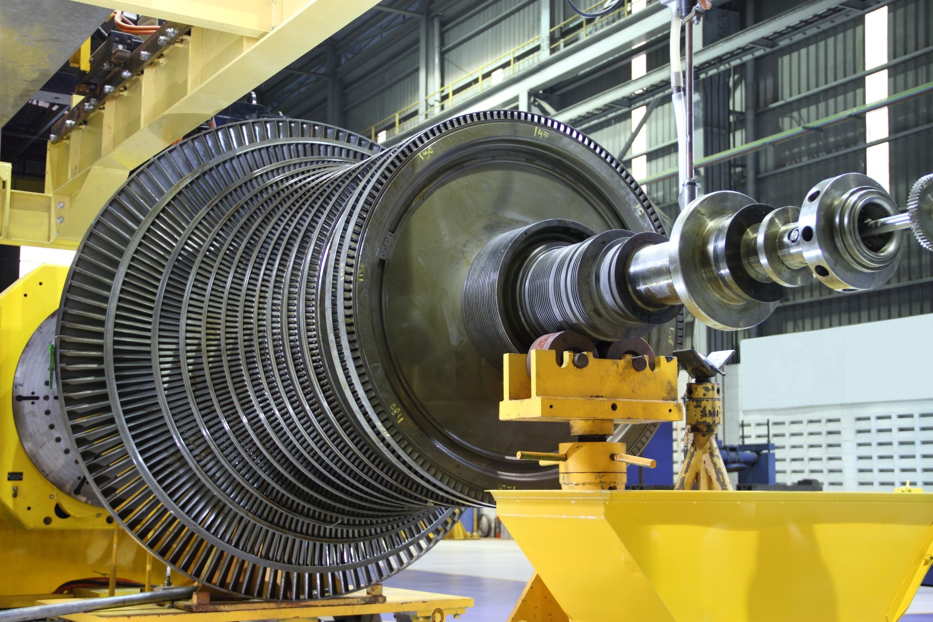 A large, multi-stage turbine rotor with numerous metal blades sits on a yellow industrial stand inside a factory.