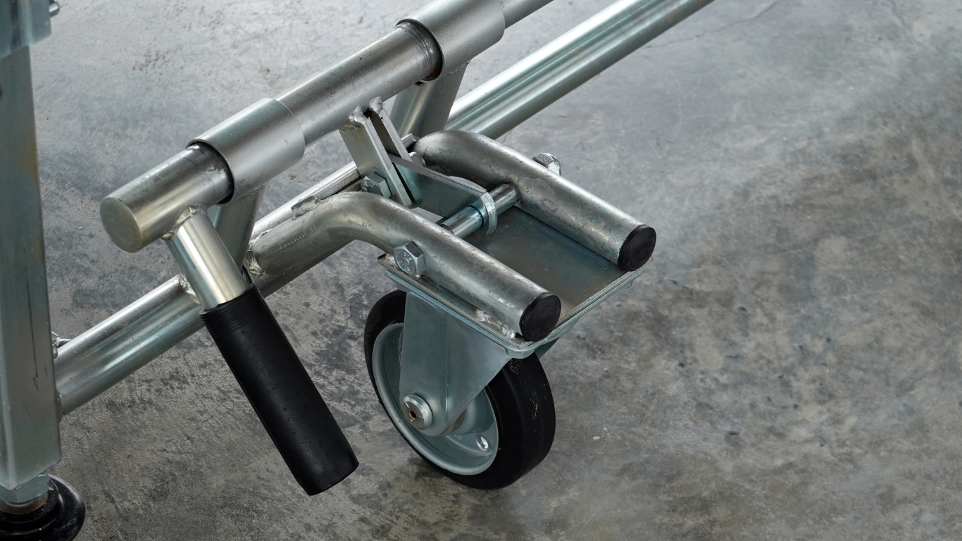 A metal caster wheel attached to a scaffolding frame with a black handle, positioned on a concrete floor.