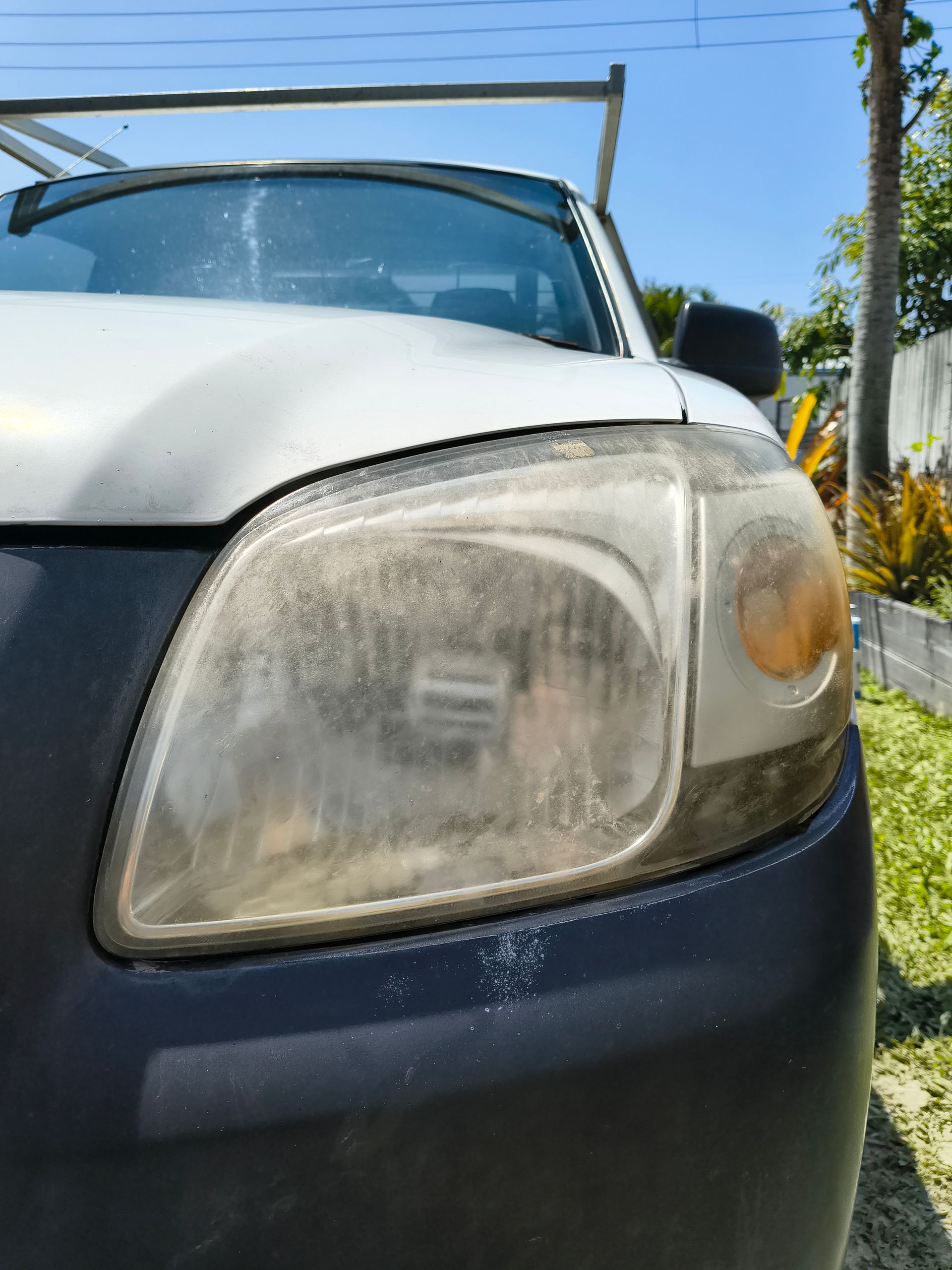 Close-up Of A White Car's Dusty Headlight. Black Bumper, Clear Sky, And Foliage Are In The Background — Best Car Detailing in Vincent, QLD