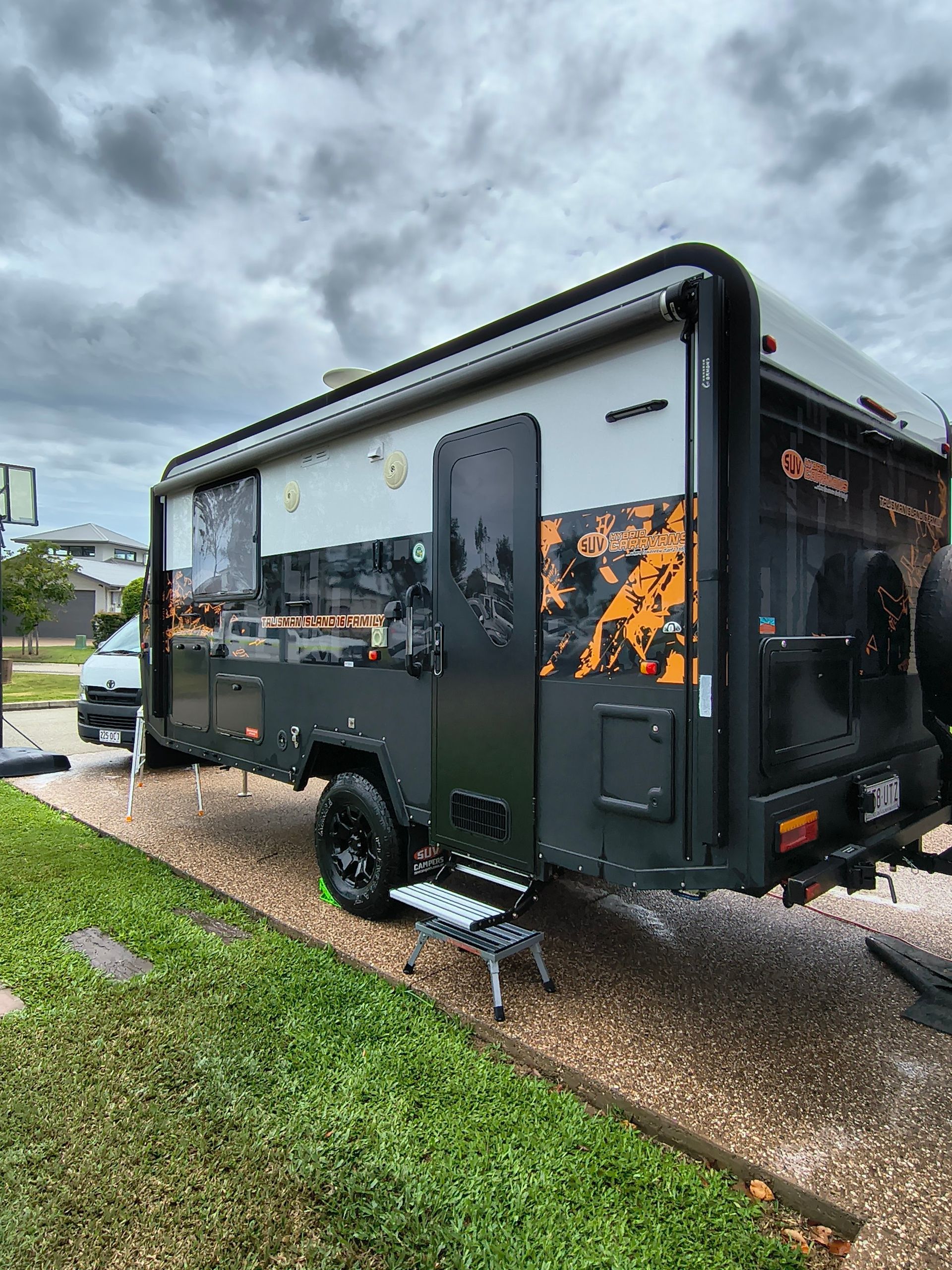 A Dark Grey And Orange Caravan Parked On Gravel With An Open Door And Steps On A Cloudy Day — Best Car Detailing in Vincent, QLD