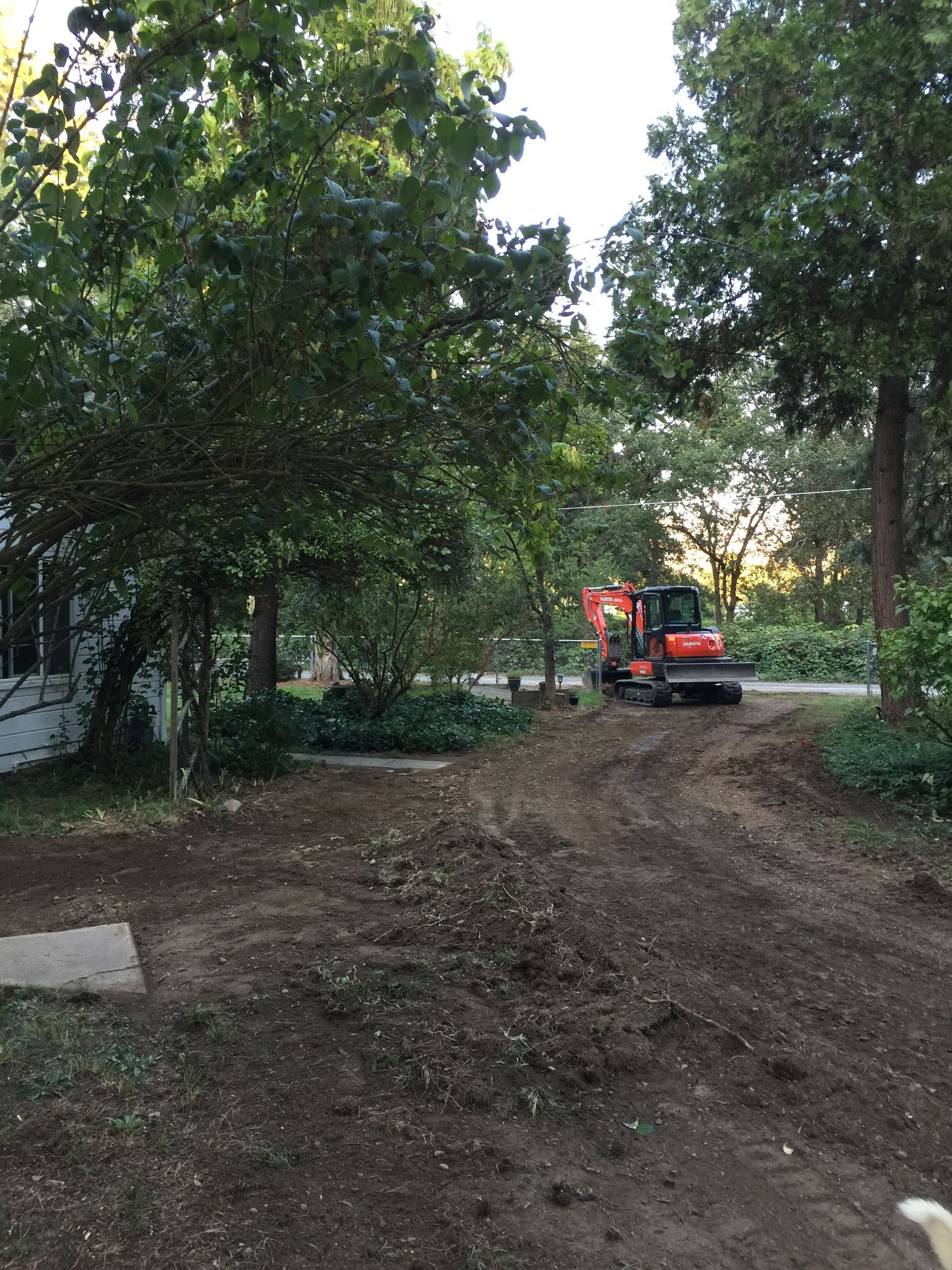 An excavator is driving down a dirt road in front of a house.
