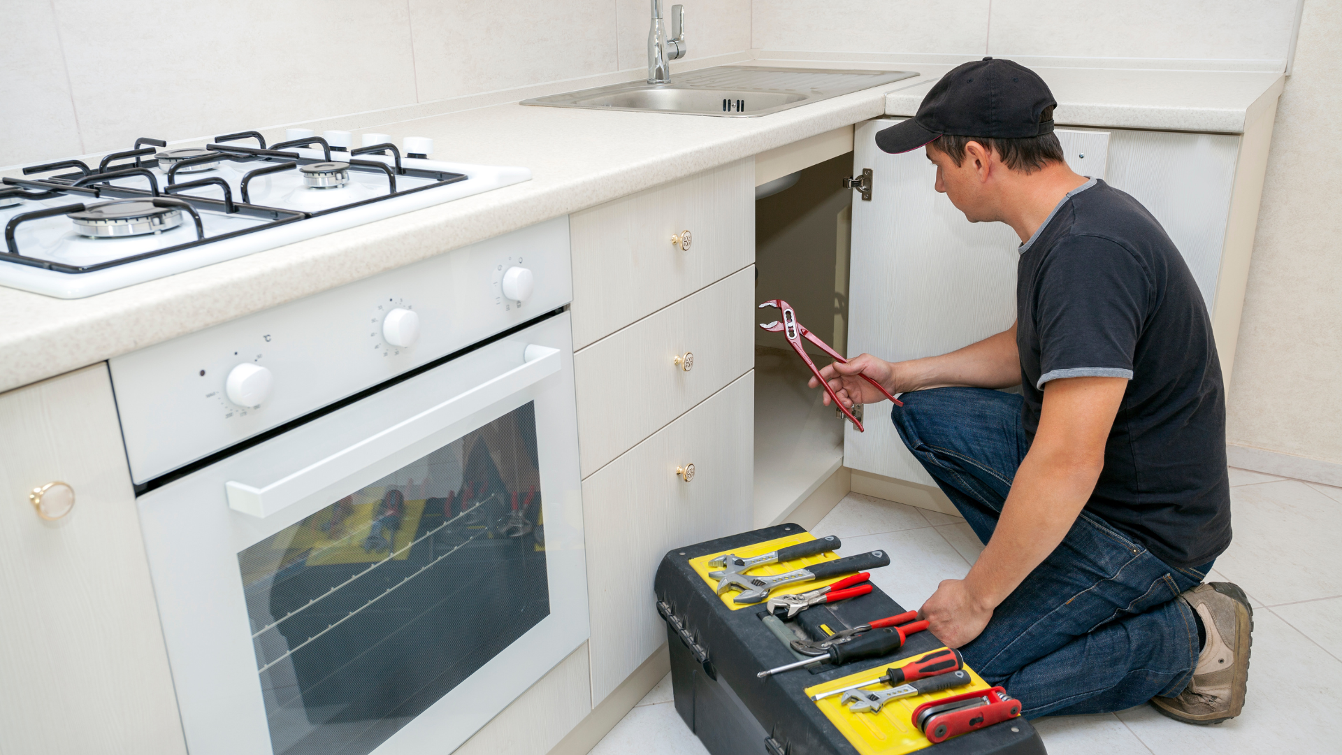A man is kneeling down in a kitchen holding a wrench.