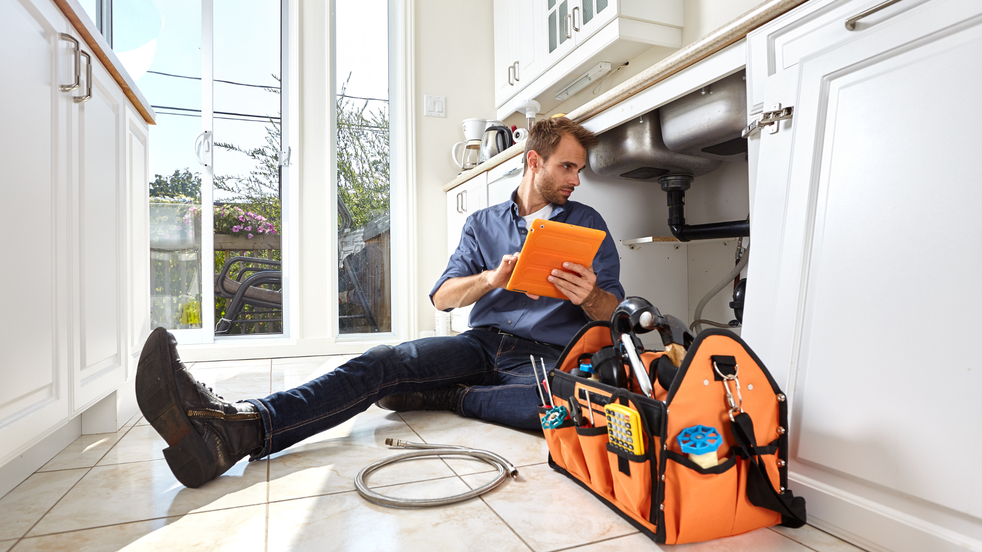 A man is sitting on the floor in a kitchen looking at a tablet.