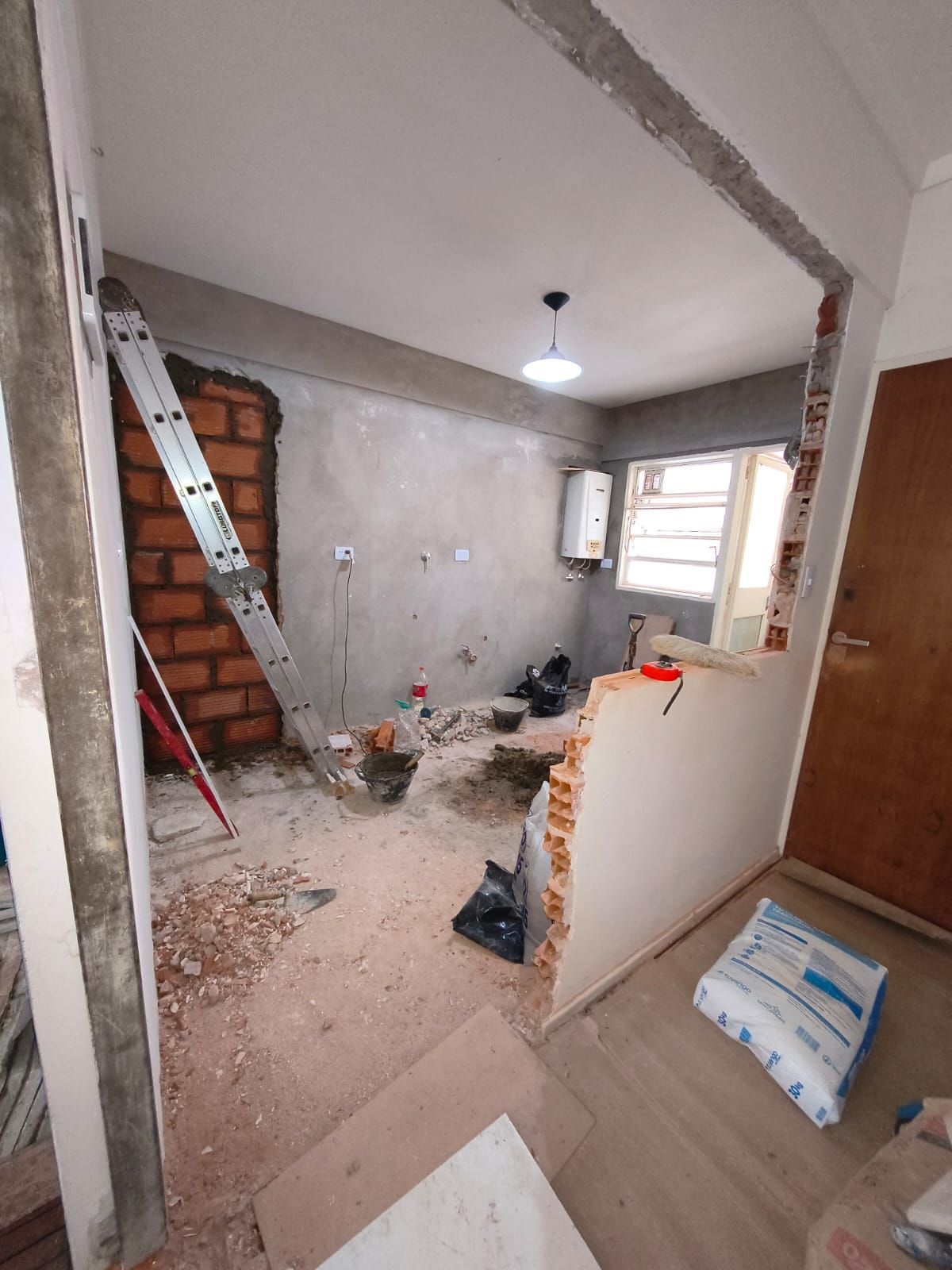Zona de reforma con ladrillo visto, escombros en el suelo, escalera y paredes sin terminar en una habitación pequeña.