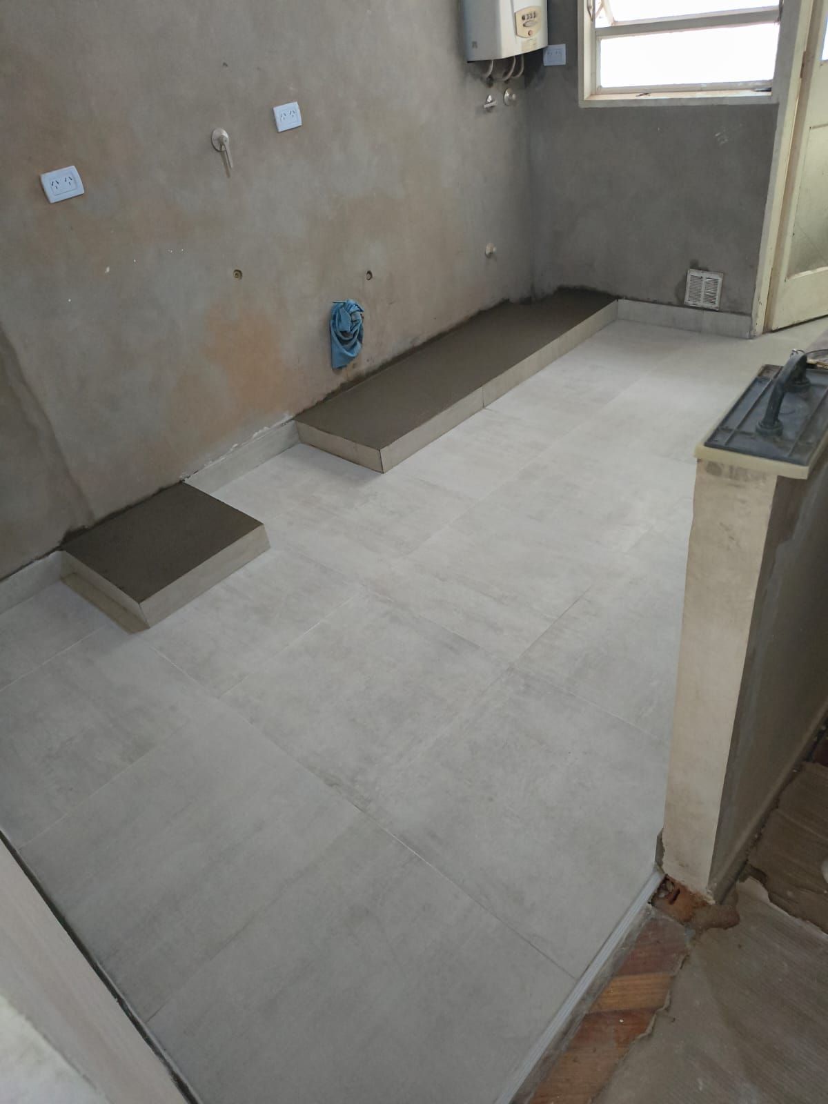 Habitación vacía con paredes sin terminar, suelo de baldosas claras y dos plataformas bajas de hormigón cerca de una ventana.