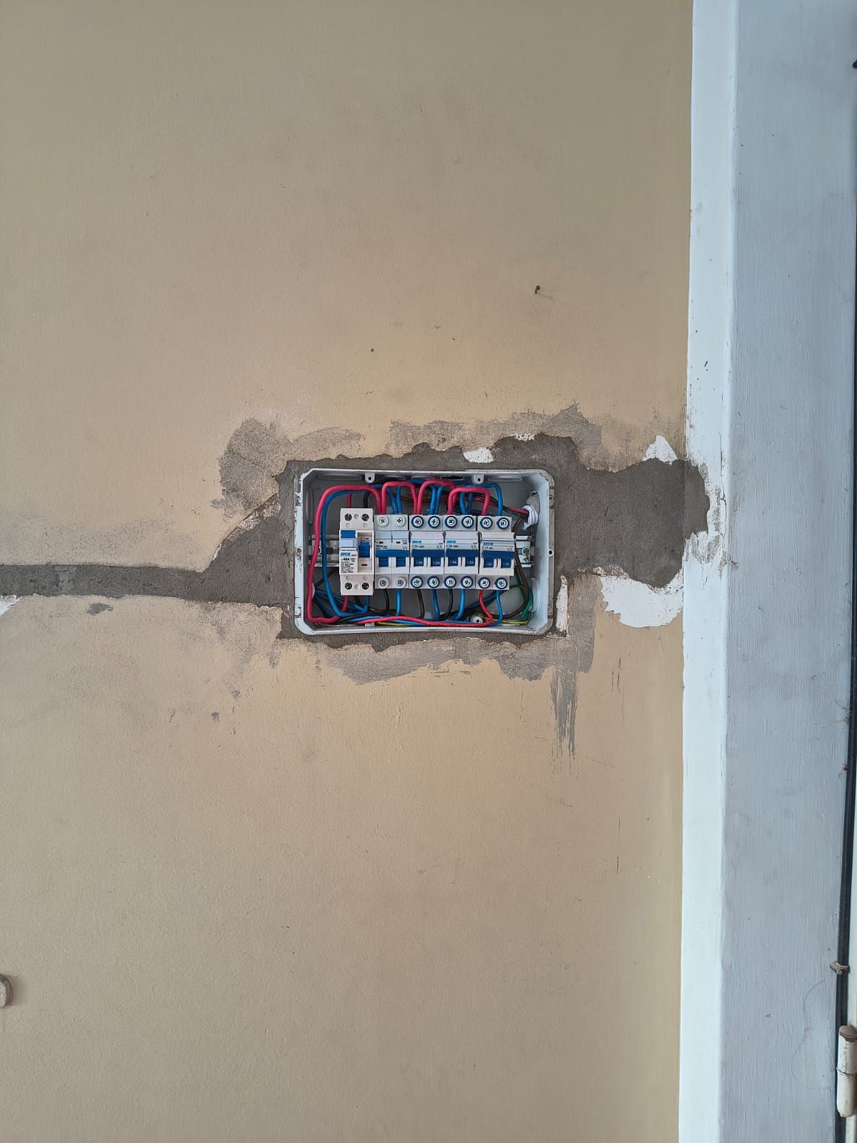 Panel de disyuntores eléctricos expuesto en un hueco de la pared con cables rojos y azules visibles.