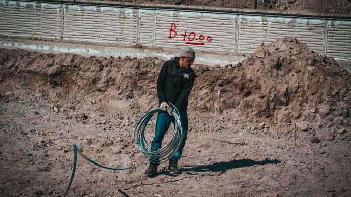 Un trabajador se encuentra de pie en una zanja de tierra, sosteniendo una manguera enrollada