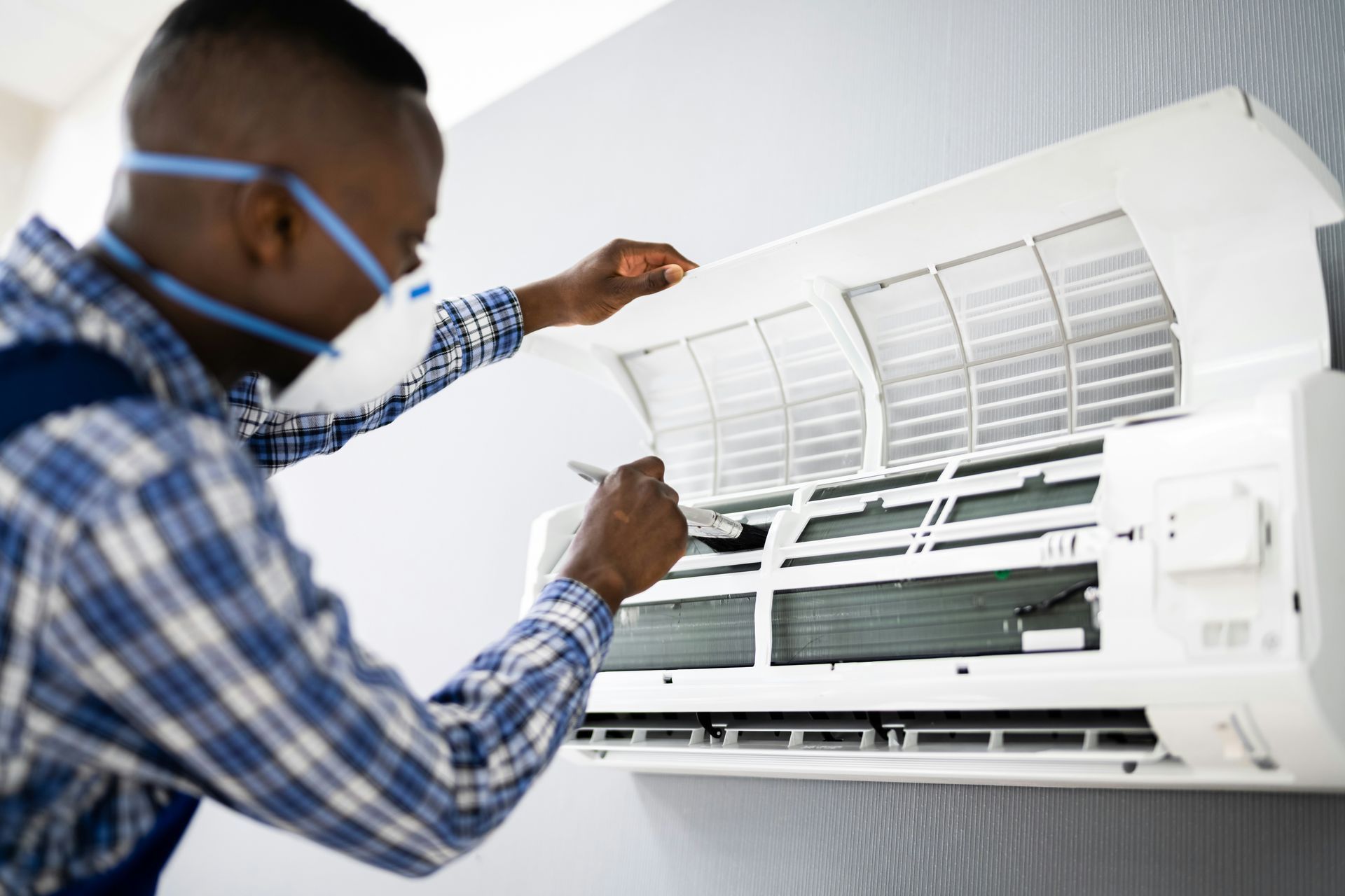 A person wearing a face mask and plaid shirt cleans the filter of a wall-mounted air conditioner.
