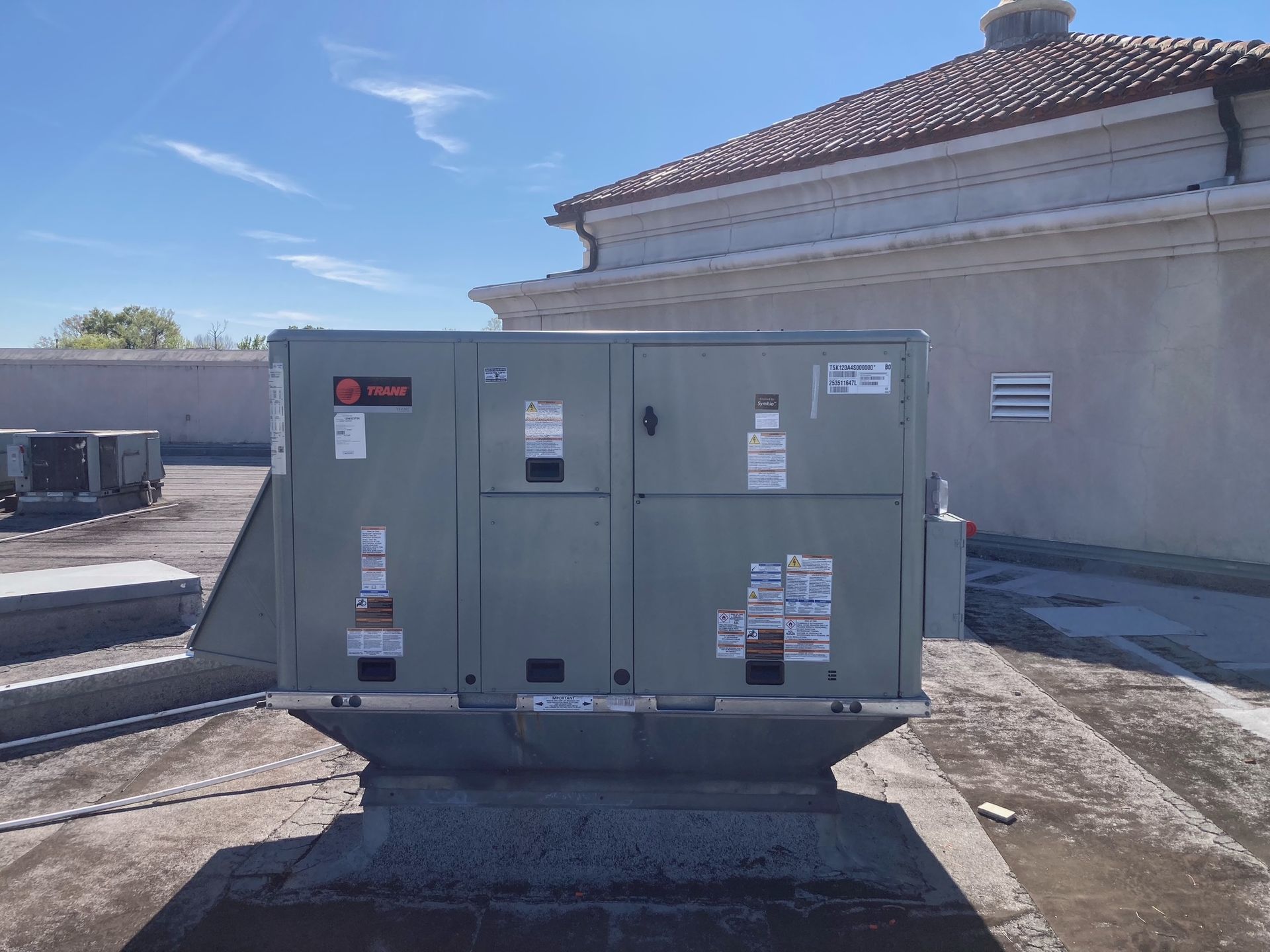 A gray Trane HVAC rooftop unit sits on a flat building roof under a bright blue sky.