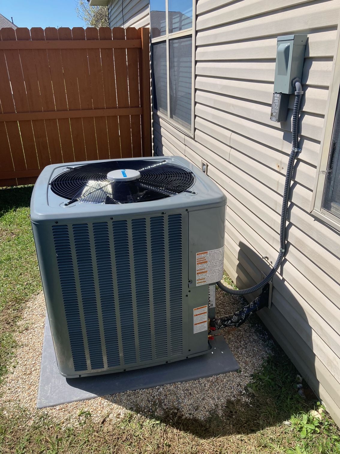 A gray outdoor air conditioning unit sits on a pad next to a beige-sided house with a wooden fence in the background.