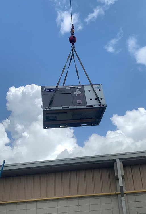 A crane lifts a large industrial HVAC unit through the air toward the roof of a commercial building.