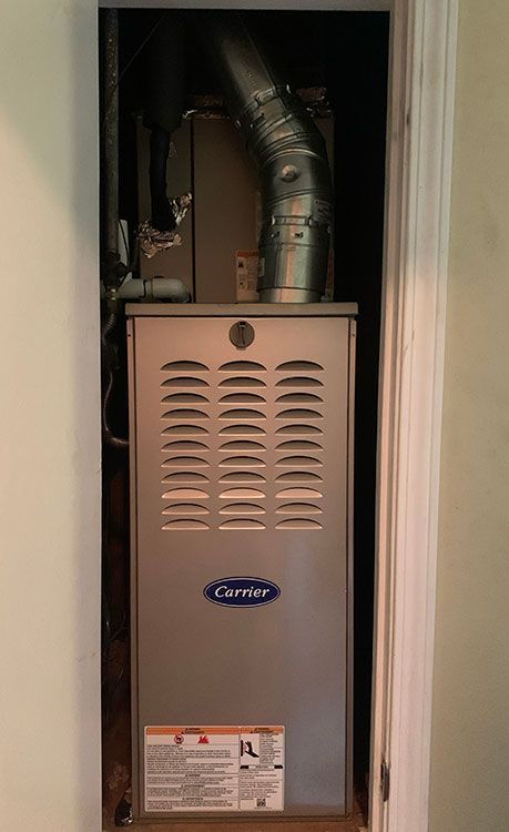 A silver Carrier HVAC furnace unit installed inside a narrow indoor closet, featuring a metal exhaust vent pipe on top.